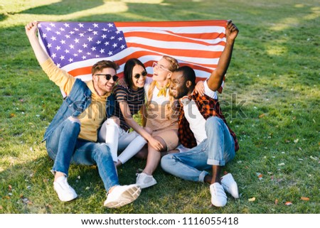 multiracial cheerful friends with american flag sitting on green grass in park