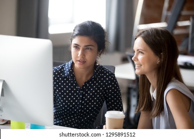 Multiracial Colleagues Indian And Caucasian Young Women Sitting Together At Desk In Office Room Sharing Ideas. Diverse Students Interns Friends Looks At Pc Screen Talking Discussing Working Moments.