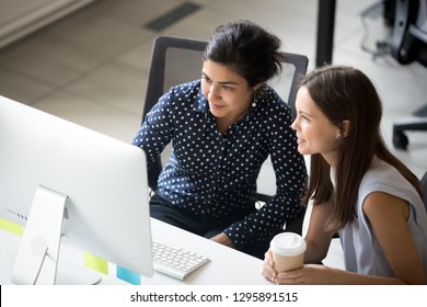 Multiracial colleagues indian and caucasian young women having coffee break sitting together at desk in office. Diverse students interns friends looks at pc screen talking discussing working moments