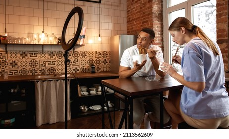 Multiracial Couple Eat Noodles During Make Eat Fast Food Video Challenge On Smartphone. Video Blogging. Black Man And Caucasian Woman Of Generation Z Create Content For Social Networks At Home Kitchen