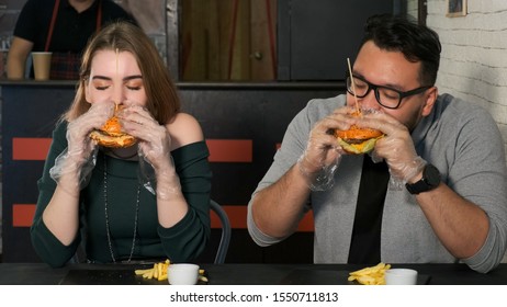 Multiracial Couple Eating Burgers In A Cafe. Asian Guy Bites A Delicious Burger