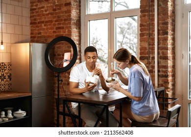 Multiracial Couple Eating Noodles At Table During Making Eat Fast Food Video Challenge On Smartphone. Video Blogging. Man And Woman Of Generation Z Creating Content For Social Networks At Home Kitchen