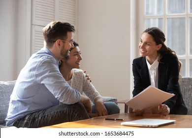 Multiracial Couple Indian Wife Caucasian Husband Hugging Feels Satisfied With Meeting Results With Realtor, Diverse People Client And Banker Seated On Sofa Discuss Mortgage Loan Contract Terms Concept