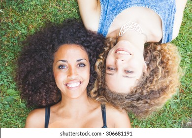 Multiracial Couple Lying On The Grass. They Are Two Young Women Resting At Park. One Is Caucasian And The Other Is Black, Both Have Curly Hair. They Are Smiling And Wearing Summer Clothes.