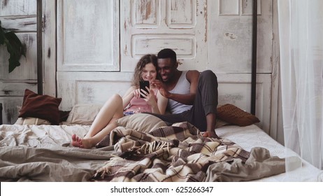 Multiracial couple in pajamas sitting on bed and use smartphone. African man and european woman looking photos, laughing - Powered by Shutterstock