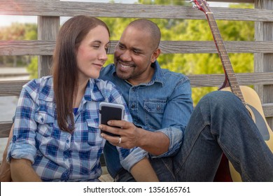  A Multiracial Couple Sits On A Deck Reviewing Selfies They Smirk At Each Other While Looking At Selfies On The Smartphone As The Sun Peaks Through The Clouds In The Background.