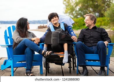 Multiracial Couple Sitting With Disabled Twelve Year Old Son In Wheelchair While Sitting In Blue Adirondack Chairs On Wooden Pier By Lake On Summer Day