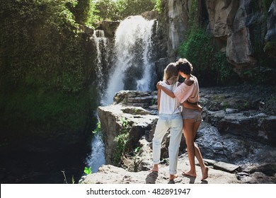 Multiracial Couple Of Two Young Girlfriends Hugging During Their Summer Vacation In Bali. Explore Waterfall In Ubud Area, Tegenungan. Gay Lovers Traveling, Discover Jungles