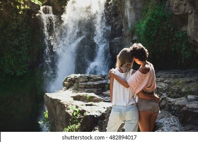 Multiracial Couple Of Two Young Girlfriends Hugging During Their Summer Vacation In Bali. Explore Waterfall In Ubud Area, Tegenungan. Gay Lovers Traveling, Discover Jungles