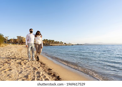 Multiracial Couple Of Young Millennials In Love Embracing Each Other While Walking By The Ocean Seaside At Sunset Or Sunrise Wearing Protective Face Mask Against Coronavirus In New Normal Vacations