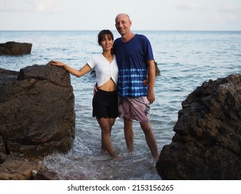 Multiracial Couple, Young Woman With Older Man Standing In The Sea Between Two Rocks On Kho Larn In Thailand  