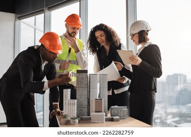 Multiracial coworkers builders and architects in formal outfit and helmets standing near table with design of buildings residential project maquette. Business people working on construction project - Powered by Shutterstock