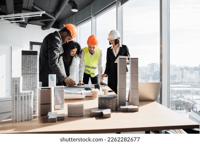 Multiracial coworkers builders and architects standing near table with blueprints, gadgets and design of buildings residential project maquette. Business people working on construction project - Powered by Shutterstock