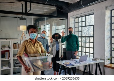 Multiracial Coworkers With Face Masks Greets Black Businesswoman Who Return Back To Work With Plastic Box Full Of Office Supplies