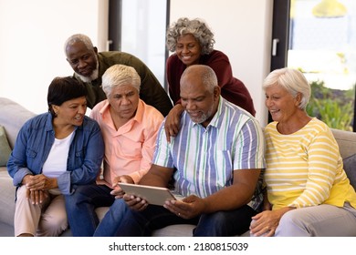 Multiracial Curious Seniors Looking At Male Friend Using Digital Tablet While Sitting On Sofa. Nursing Home, Wireless Technology, Unaltered, Togetherness, Support, Assisted Living, Retirement.