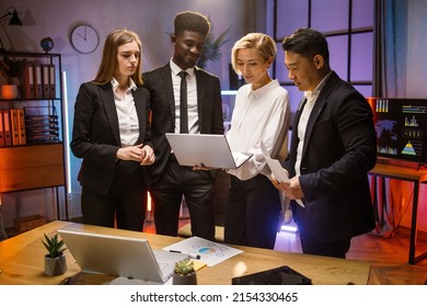 Multiracial Diverse Work Group Standing At The Table Brainstorming, Using Laptop And Project Requirements To Develop Startup Strategy Plan. Four Colleagues Brainstorming At Dark Office