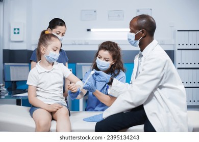 Multiracial doctor and nurse in consultation room caring for young patient and her mother. Male physician and female medical assistant wrapping sterilized bandage on the arm of a little girl. - Shutterstock ID 2395134243