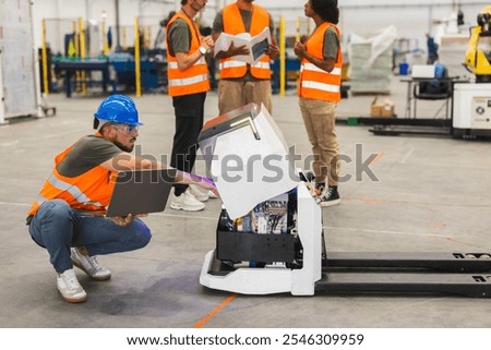 Multiracial engineering team working together, maintaining an automated guided vehicle - agv - in a large industrial factory, using laptop and documents