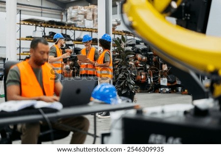 Multiracial engineers collaborating in high tech factory with robotic arms, using laptop for planning, showcasing teamwork and tech advancement in automation