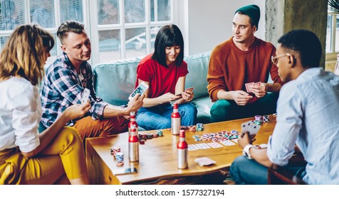 Multiracial Enthusiastic Concentrated Team Holding Cards While Playing Poker Game Around Table And Drinking Beverages During Spending Excellent Time Together At Home