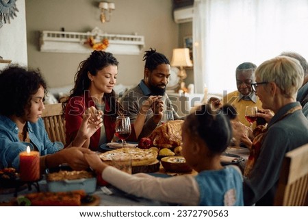 Multiracial extended family saying grace and holding hands while gathering for Thanksgiving meal at dining table.