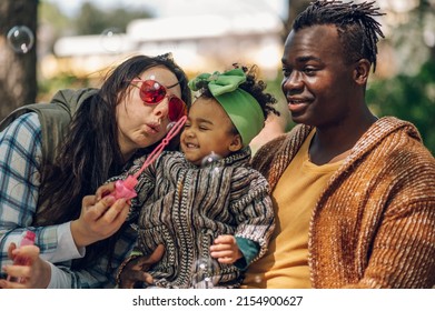 Multiracial Family Concept. Black Father And White Mother Playing With Their Daughter In The Park And Spending The Day Together. Blowing Soap Bubbles.