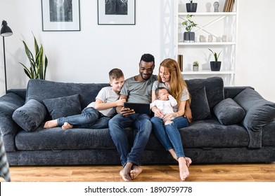 A multiracial family relaxing with a digital tablet sitting on the comfortable sofa, watching video, playing game on the touchscreen, have fun together. Spouses, baby girl and school-age son - Powered by Shutterstock