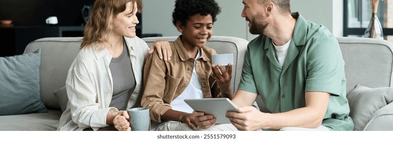 Multiracial family sitting on a couch, sharing a happy moment while using a tablet and drinking hot beverages. Parents enjoying quality time with their smiling son, who is holding a smartphone - Powered by Shutterstock