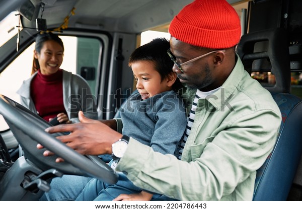 A multiracial father teaches his son how\
to drive a van and explains to him how it\'s working while a mother\
in the background smiles and looks at\
them.