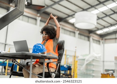 Multiracial female engineer in a robotics factory takes a break, stretching while working on a laptop in a bright, modern workspace filled with industrial equipment - Powered by Shutterstock