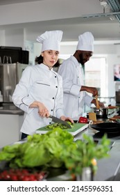 Multiracial Food Industry Workers Standing In Restaurant Professional Kitchen While Cooking Tasty Dish. Confident Head Chef Cutting Fresh Vegetables For Garnish While Smiling At Camera.