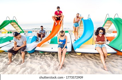 Multiracial friends with closed face mask using tracking app with mobile smart phone - Bored young milenial people sitting at beach - New normal lifestyle and social distancing concept - Bright filter