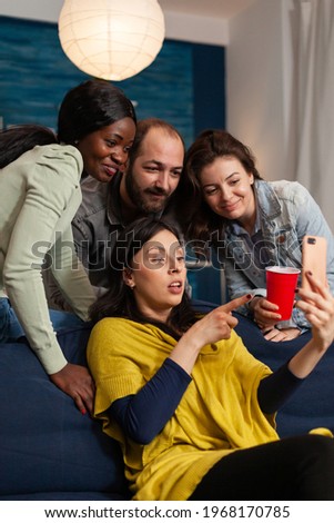 Multiracial friends hanging out late at night watching funny entertainment video on smartphone. Group of multiracial people spending time together sitting on couch late at night in living room.