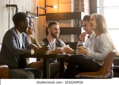 Multiracial friends having fun and laughing drinking coffee in coffeehouse, diverse young people talking joking sitting together at cafe table, multi ethnic millennials spending time in coffee shop