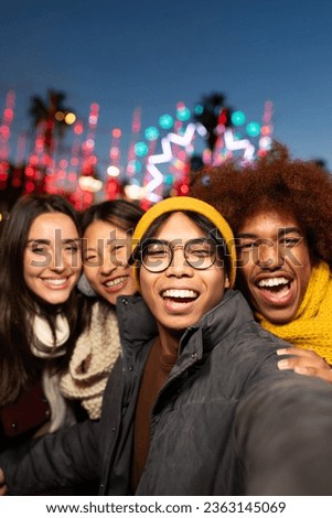 Multiracial friends having fun together taking vertical selfie during winter market at night looking at camera.