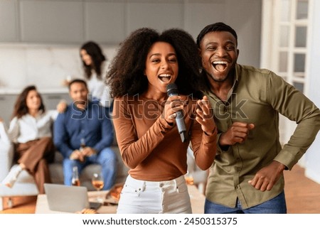 Multiracial friends share a microphone, singing joyously with laughter and fun expressions in a cozy home setting