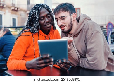 Multiracial friends sharing digital content on tablet computer while sitting at outdoor cafe. African woman in orange hoodie and Caucasian man collaborating in urban setting. - Powered by Shutterstock