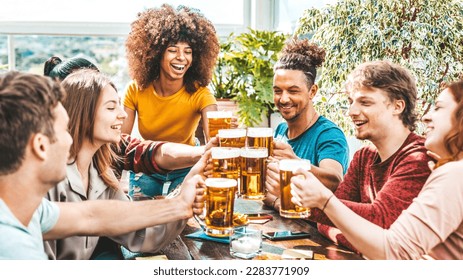 Multiracial friends toasting beer glasses at brewery pub garden - Happy young people enjoying happy hour sitting at bar restaurant - Friendship concept with guys and girls drinking and eating together - Powered by Shutterstock