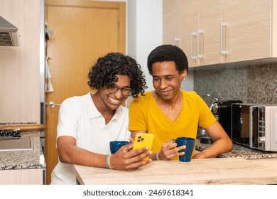 Multiracial gay couple sitting on the kitchen enjoying coffee cup and using phone - Powered by Shutterstock