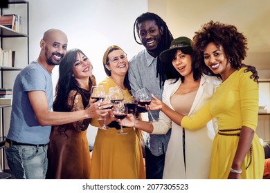 Multi-racial Group Of Adults Smiling And Looking At The Camera Holding Red Wine Glasses - Multiracial Friends Standing Not He Living Room An Toasting Red Wine