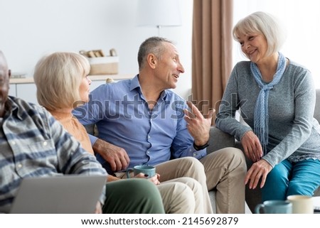 Multiracial group of cheerful pensioners drinking tea and chatting, happy senior women and men in casual sitting on couch in living room, enjoying time together at sanatorium, having conversation