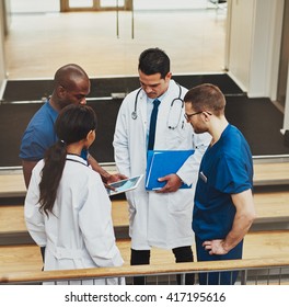 Multiracial Group Of Doctors In An Impromptu Meeting Standing On A Stairwell Looking At Information On A Tablet Computer, View From Above