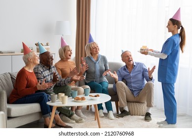 Multiracial group of excited elderly people men and women wearing colorful paper hats having birthday party at nursing home, young woman nurse holding birthday cake with lit candles, copy space - Powered by Shutterstock