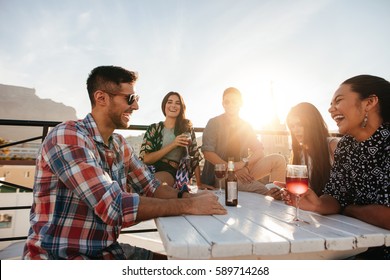 Multiracial Group Of Friends Having Cocktail Party On The Rooftop. Young Men And Women Sitting Around Table With Drinks And Laughing.