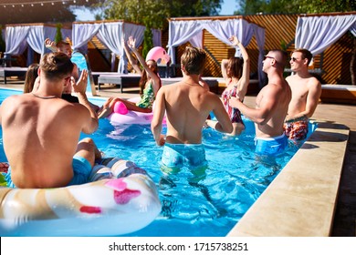 Multiracial Group Of Friends Having Party In A Private Villa Swimming Pool. Happy Young People In Swimwear Dancing And Clubbing With Inflatable Flamingo, Swan, Floaty In Luxury Resort On Sunny Day.