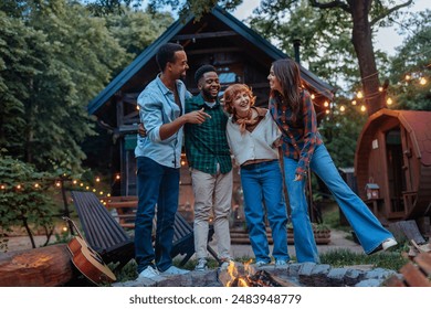 A multiracial group of friends standing and laughing next to a burning fire pit in the backyard of a cozy cabin in the woods. - Powered by Shutterstock