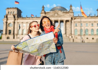 Multiracial Group Of Friends Visiting The City Of Berlin. Two Women Reading Map With Reichstag Building On The Background. Friendship And Travel Concept With Real Candid Emotions