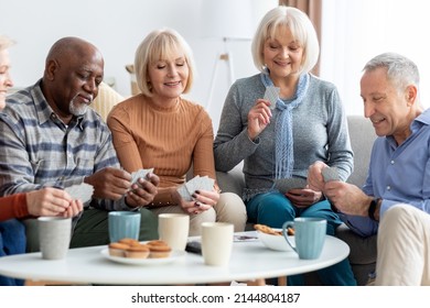 Multiracial group of happy senior people sitting on couch, playing cards, drinking tea, eating cookies at nursing home, enjoying time together. Residential facilities, assisted living concept - Powered by Shutterstock