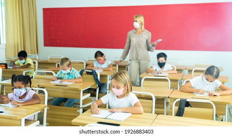 Multiracial Group Of Kids Wearing Face Masks Working At Class, Writing And Listening Explanations Of Teacher In Classroom