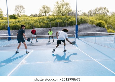 A multiracial group of players engages in a doubles pickleball match on a blue court during a sunny day. Concept of teamwork and diversity in sports. - Powered by Shutterstock